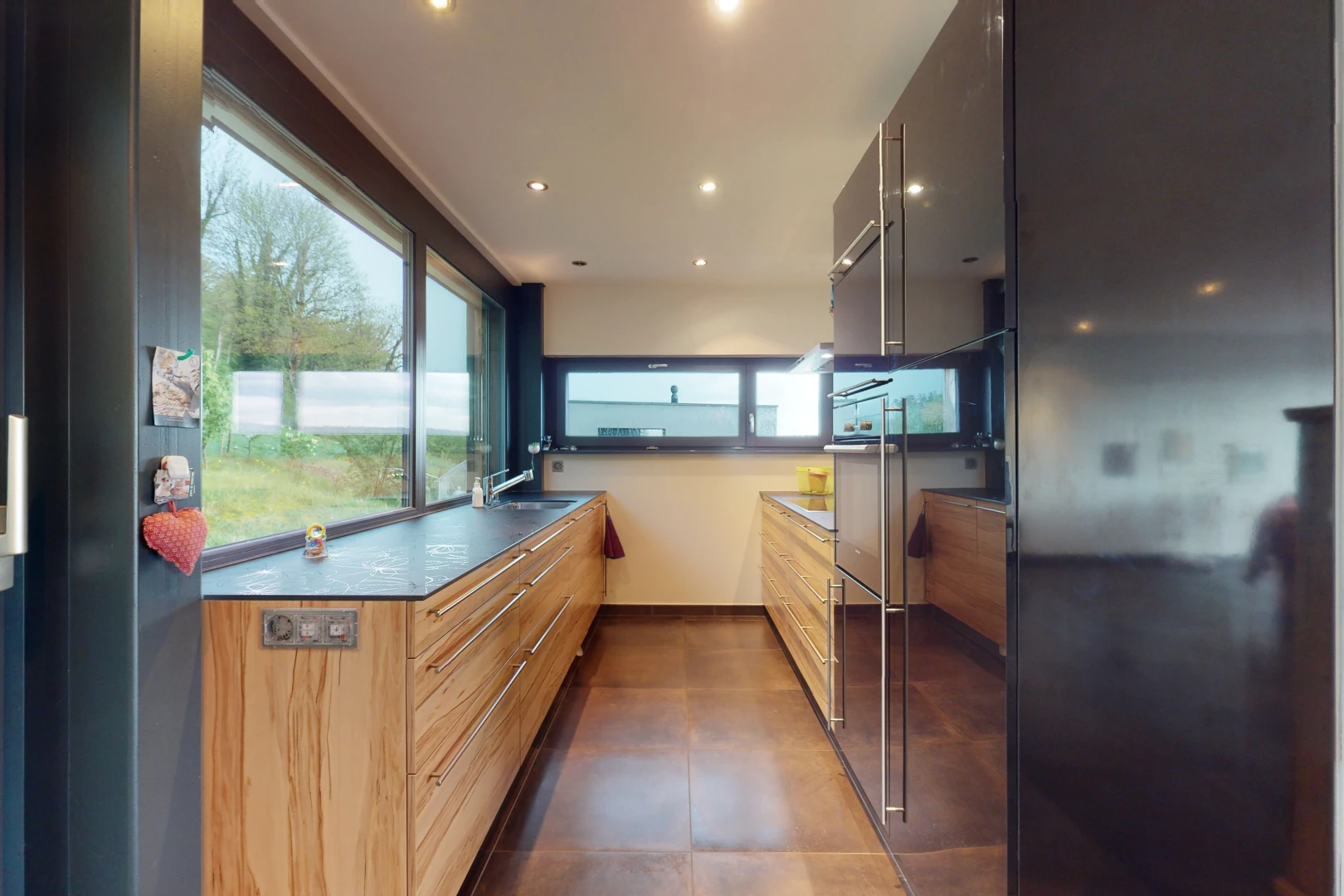 cuisine avec lumière naturelle, carrelage, sol foncé, plan de travail foncé, et meubles marron
