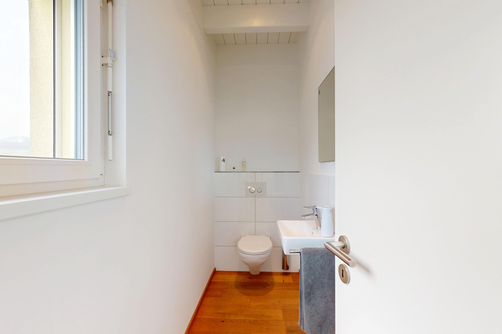 salle de bains avec lumière naturelle, miroir, sol en finition bois, et toilette