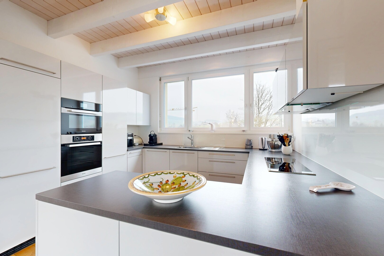 cuisine avec plaque de cuisson, hotte, plafond à poutres, armoires blanches, et lumière naturelle