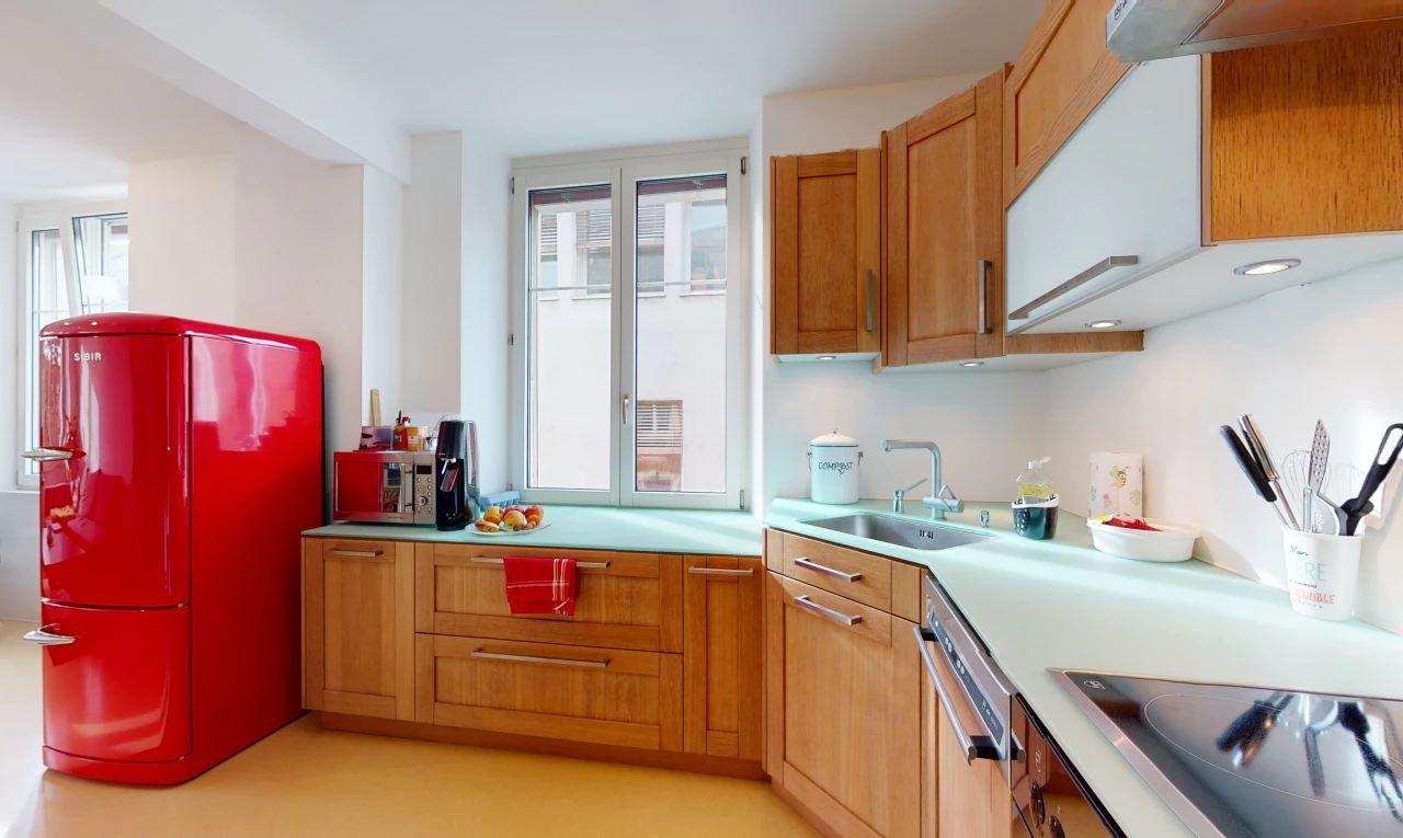 cuisine avec lumière naturelle, poutres apparentes, cuisinière électrique, lave-vaisselle, réfrigérateur, plan de travail clair, sol clair, et meubles marron