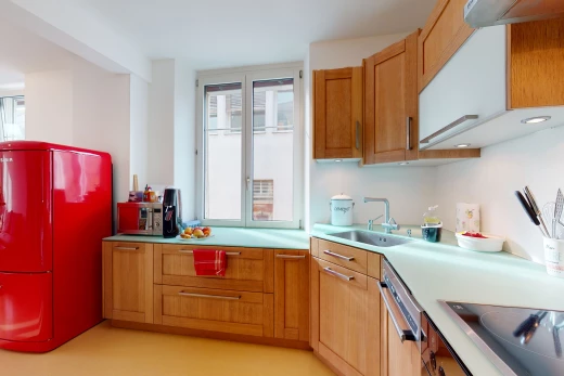cuisine avec lumière naturelle, poutres apparentes, cuisinière électrique, lave-vaisselle, réfrigérateur, plan de travail clair, sol clair, et meubles marron