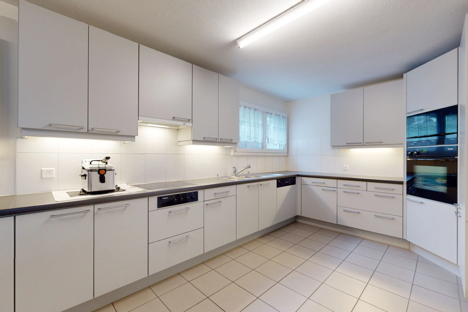 cuisine avec lumière naturelle, carrelage, four, cuisinière électrique, sol clair, et meubles blancs