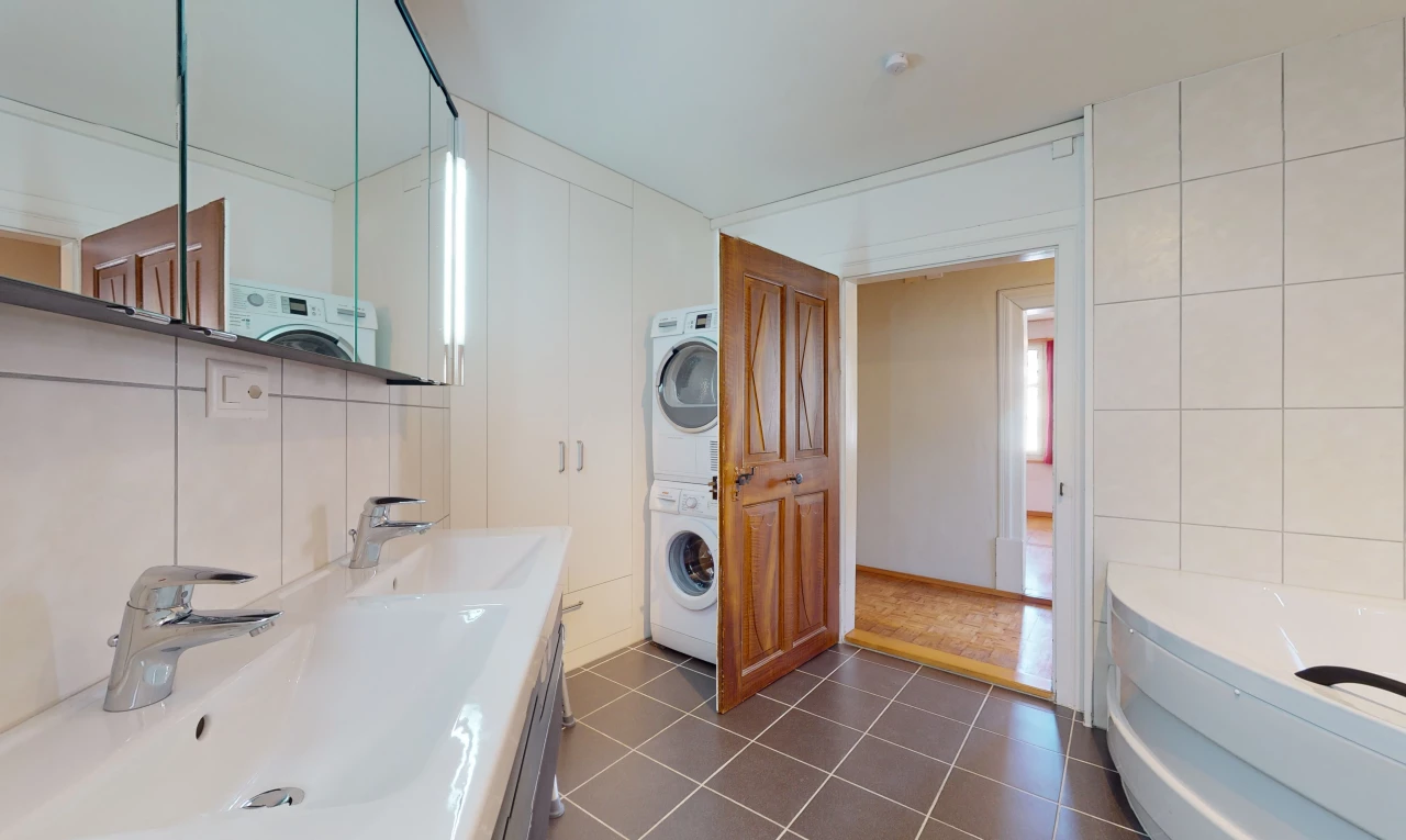 salle de bains avec carrelage, machine à laver / sèche-linge, meuble-lavabo, baignoire, double vasque, et miroir